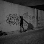 Een persoon in donkere kleding loopt 's nachts alleen langs een met graffiti bedekte betonnen muur langs een verlaten straat. De scène is zwak verlicht en creëert een stemmige, stedelijke sfeer.