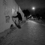 Een persoon in donkere kleding spuit 's nachts graffiti op een muur langs een leeg, geplaveid pad dat wordt verlicht door straatlantaarns.