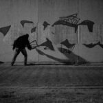 Een persoon in donkere kleding spuit 's nachts graffiti op een grote betonnen muur en werpt een lange schaduw. De scène is zwak verlicht en een rugzak rust op de grond vlakbij.