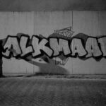 Een persoon spuit 's nachts grote graffitiletters met de tekst "ALKMAAR" op een betonnen muur. De scène is in zwart-wit en de schaduw van de persoon verschijnt op de muur.