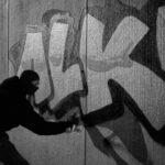 Een persoon met capuchon en masker spuit 's nachts grote graffitiletters op een betonnen muur. De afbeelding is zwart-wit en de letters "ALK" zijn prominent aanwezig.