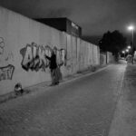Iemand spuit 's nachts graffiti op een betonnen muur langs een verlaten, slecht verlichte straat. Op de muur staan kleurrijke ontwerpen, waaronder een lachende bloem en de tekst "MTV.