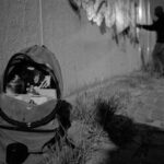 Een zwart-wit foto toont een rugzak met spuitbussen op de grond naast een muur. Op de achtergrond spuit iemand 's nachts graffiti op de muur onder straatverlichting.