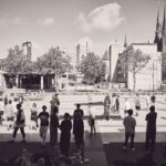 Een groep mensen verzamelt zich op een open stedelijk plein met bomen, moderne gebouwen en een kerk met hoge torenspitsen op de achtergrond op een zonnige dag. De afbeelding is in zwart-wit.