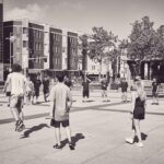 Een groep mensen staat in een grote cirkel buiten op een stadsplein, sommigen lijken een spel te spelen of te sporten. Gebouwen, bomen en voetgangers zijn zichtbaar op de achtergrond. De afbeelding is in zwart-wit.