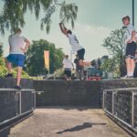Vier jonge mannen in sportkleding oefenen parkour buiten; één springt in de lucht over een spleet tussen twee stenen muren terwijl de anderen toekijken. Bomen en een bus zijn zichtbaar op de zonnige achtergrond.