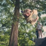Een persoon in een bruin shirt voert een dynamische salto of sprong uit bij een boom in een park, terwijl drie anderen toekijken vanaf de grond, twee staand en één zittend op een stenen constructie. Zonlicht filtert door de bladeren.