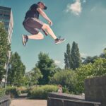Een man in atletische kleding springt in de lucht tussen stenen muren, terwijl een andere man vlakbij staat toe te kijken. Bomen en groen omringen hen onder een gedeeltelijk bewolkte hemel.