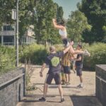 Vier mensen oefenen acrobatiek in de buitenlucht; één persoon wordt in de lucht getild door een ander terwijl twee spotters in de buurt staan. Bomen en een modern gebouw zijn zichtbaar op de achtergrond op een zonnige dag.