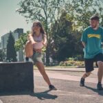 Een jonge vrouw in korte broek en tanktop springt over een laag betonnen muurtje terwijl een jonge man in een blauw T-shirt haar glimlachend volgt. Ze zijn buiten op een zonnige dag met bomen en gebouwen op de achtergrond.