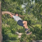 Een man in een grijs t-shirt en een zwarte korte broek hangt met beide armen en gebogen benen aan een boomtak, omringd door weelderig groen gebladerte in een zonovergoten buitenomgeving.