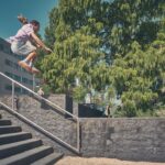 Een persoon in atletische kleding springt in de lucht over een metalen reling en stenen treden in een stedelijk buitengebied, met groene bomen en gebouwen op de achtergrond onder een heldere hemel.
