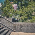 Een persoon wordt midden in de lucht vastgelegd terwijl hij een backflip maakt over een metalen leuning naast een buitentrap, met bomen en gebouwen op de achtergrond onder een zonnige hemel.