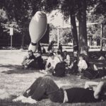 Een groep mensen ontspant op het gras in een park, sommigen zittend en sommigen liggend, bij een grote opblaasbare bowlingpin. Bomen zorgen voor schaduw en een paar mensen staan of lopen op de achtergrond. De afbeelding is in zwart-wit.