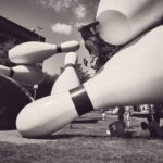 Een persoon klimt op gigantische bowlingpinsculpturen in een park terwijl anderen in het gras vlakbij zitten; de scène is in zwart-wit.