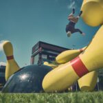 Een persoon flipt in de lucht tussen grote opblaasbare gele bowlingkegels en een zwarte bowlingbal, terwijl anderen in de buurt staan of toekijken op een grasveld met gebouwen op de achtergrond.