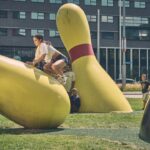 Een jonge vrouw skateboardt over een grote, gele bowlingpinsculptuur in een stadspark terwijl mensen op de achtergrond toekijken.