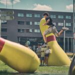 Een persoon springt in de lucht tussen twee grote, gele bowlingpinsculpturen in een stedelijk buitengebied, terwijl drie mensen in de buurt toekijken. Op de achtergrond zijn een modern gebouw en straatnaamborden te zien.