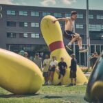 Een shirtloze man springt over een grote, zwarte bowlingbal, met oversized bowlingpinsculpturen en een groep mensen op de achtergrond op een zonnige dag.