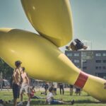 Verschillende jongeren verzamelen zich bij grote gele bowlingpinsculpturen in een graspark. Eén persoon is in de lucht en maakt een salto van het beeld, terwijl anderen toekijken of in de buurt ontspannen. De lucht is meestal helder.
