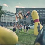 Een jongeman maakt een backflip in de lucht op een grasveld met grote, kleurrijke bowlingpinsculpturen, terwijl een groep mensen op de achtergrond toekijkt bij stadsgebouwen en geparkeerde auto's.