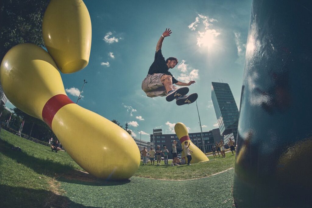 Een persoon in de lucht voert een truc uit boven een grote gele bowlingpin sculptuur buiten, met een menigte en moderne gebouwen op de achtergrond onder een zonnige hemel.