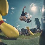 Een persoon in de lucht voert een truc uit boven een grote gele bowlingpin sculptuur buiten, met een menigte en moderne gebouwen op de achtergrond onder een zonnige hemel.