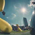 Een persoon flipt in de lucht in de buurt van gigantische gele bowlingpinsculpturen op een grasveld, met verschillende mensen in de buurt, stadsgebouwen op de achtergrond en de zon die schijnt in een blauwe lucht.