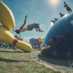 Een persoon maakt een backflip in de lucht in de buurt van grote, gele bowlingpinsculpturen buiten, terwijl mensen toekijken en de lucht zonnig is.