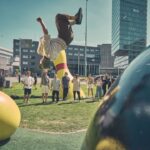 Een persoon maakt een backflip in een stadspark, omringd door grote gele sculpturen en een groep toeschouwers, met hoge moderne gebouwen op de achtergrond.