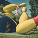 Een shirtloze man in korte broek en pet springt tussen reusachtige gele bowlingkegels naast een grote zwarte bowlingbal in een openluchtpark op een zonnige dag.