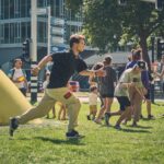 Een jonge man rent op gras in een levendig buitengebied, omringd door mensen die staan, wandelen en praten. Op de achtergrond zijn grote gele kegelstructuren en stadsgebouwen te zien.