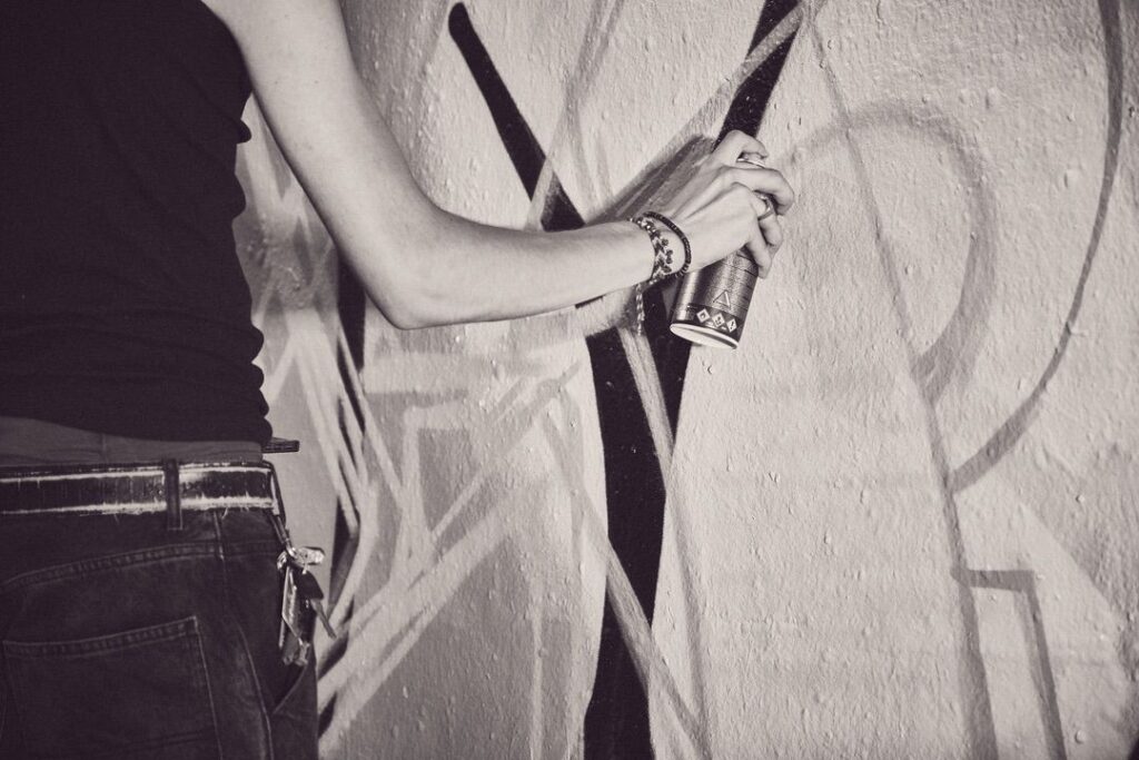 Een persoon in spijkerbroek en mouwloos topje spuit zwarte lijnen op een muur bedekt met graffiti, te zien vanaf het middel met gestrekte arm. De afbeelding is zwart-wit.