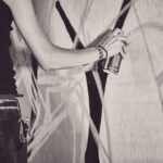 Een persoon in spijkerbroek en mouwloos topje spuit zwarte lijnen op een muur bedekt met graffiti, te zien vanaf het middel met gestrekte arm. De afbeelding is zwart-wit.