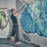 Een persoon met een zwarte tanktop, donkere spijkerbroek en pet spuit graffiti op een grote muur bedekt met kleurrijke tags en ontwerpen. Vlakbij ligt een beige tas op de grond.