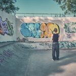 Een persoon in een zwarte tanktop en baggy jeans staat in een skatepark, tegenover een muur bedekt met kleurrijke graffitikunst onder een helderblauwe hemel en omringd door bomen.