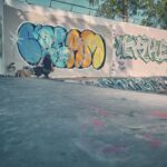 Een persoon met een hoed op zijn hurken spuit kleurrijke graffiti op de muur van een leeg skatepark onder een heldere hemel, met bomen zichtbaar boven de muur.