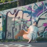 Een persoon in een lichtblauwe outfit en een pet zit gehurkt en schildert kleurrijke graffitikunst op een betonnen buitenmuur in de buurt van een vuilnisbak, met bomen zichtbaar op de achtergrond.