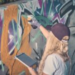 Een persoon met lang blond haar die een pet en handschoenen draagt, spuit kleurrijke graffiti op een muur terwijl hij een tablet in zijn andere hand houdt.