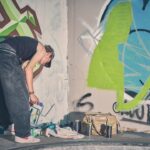 Een persoon in een wijde spijkerbroek en een tanktop buigt voorover bij een met graffiti bedekte muur en houdt een verfbus vast. Naast hem staan verfspuitbussen en een beige tas op de grond. Op de muur staat kleurrijke straatkunst.