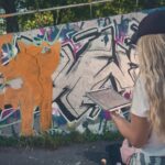 Een vrouw met lang blond haar, met een pet op en een wit T-shirt aan, staat buiten en houdt een tablet vast voor een kleurrijke muur bedekt met graffiti. Bomen en zonlicht zijn zichtbaar op de achtergrond.