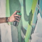 Een hand met kleurrijke geweven armbanden spuit groene verf uit een bus op een muur bedekt met abstracte graffitikunst in blauw-, groen- en wittinten.