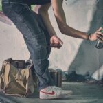 Een persoon spuit graffiti op een muur, met een groene spuitbus in de ene hand en een telefoon in de andere, met een beige tas en meer spuitbussen op de grond naast hem.