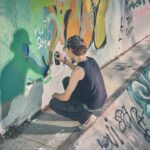 Een persoon met een zwarte tankdop en pet knielt op een betonnen oppervlak terwijl hij kleurrijke graffiti op een muur spuit, waardoor er een schaduw valt op de levendige muurschildering.