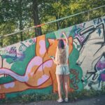 Een persoon met lang blond haar, een korte broek en een pet schildert een grote oranje kat op een met graffiti bedekte muur buiten, omringd door bomen en groen.