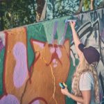 Een persoon met lang blond haar, een pet op en spuitverf in de hand, schildert buiten een kleurrijke muurschildering van een oranje kat op een met graffiti bedekte muur.