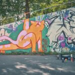 Een kleurrijke graffiti muurschildering op een buitenmuur toont een grote oranje kat die zich uitstrekt tegen een groene achtergrond. Spuitbussen en doppen liggen verspreid op de grond onder het kunstwerk. Bomen zijn zichtbaar achter de muur.