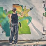 Een persoon met een pet op, een zwarte tanktop en een wijde spijkerbroek spuit kleurrijke graffiti op een witte betonnen muur, terwijl hij met zijn rug naar de camera staat. Zonlicht werpt schaduwen op de scène.
