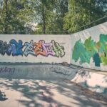 Een persoon in donkere kleding staat in een lege kom van een skatepark en kijkt naar kleurrijke graffiti op de omringende witte muren, met bomen en zonlicht op de achtergrond.