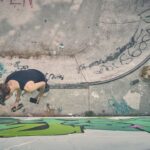 Een persoon met een zwarte tankdop en hoed spuit graffiti op een betonnen muur van een skatepark. Verfblikken, een tas en kunstbenodigdheden liggen verspreid in de buurt. De scène wordt van bovenaf bekeken.